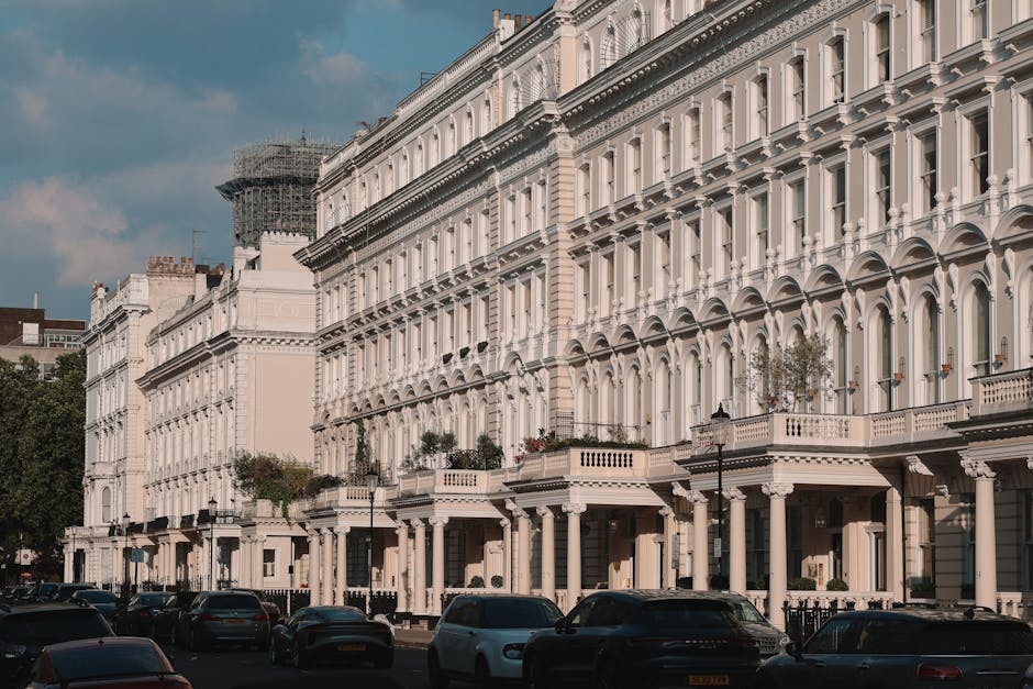 The image depicts a row of elegant, white Victorian-style terraced houses along a street, featuring ornate architectural details such as decorative cornices, arched windows, and small balconies with potted plants. In the foreground, several parked cars line the street, with a clear view of the pavement and the main building entrances. The scene is well-lit with natural daylight, and a partly cloudy sky is visible above. In the background, scaffolding surrounds a building under renovation. This setting reflects an urban residential area, likely in Earls Court, with property interiors possibly being prepared for a house move. As part of professional home relocation services, Man and a Van Earls Court may handle packing materials, furniture transport, and loading processes for these Victorian flats, utilizing vans parked nearby and equipment such as trolleys and moving blankets to facilitate the logistics of your removals in SW5.