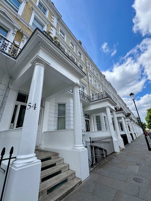 A row of white Victorian-style terraced houses with decorative columns supporting small front porches, each with a set of steps leading up from the pavement. The buildings feature large sash windows, black wrought iron railings, and number 54 displayed on the front column of one property. The scene is captured during daytime with a partly cloudy sky overhead, and a streetlamp visible along the sidewalk. The image portrays a residential area in Earls Court, suitable for house removals or furniture transport services. Occasionally, Man and a Van Earls Court provides assistance with packing and moving, often involving loading and unloading of furniture and boxes in such locations.