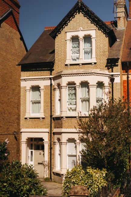 The image depicts a row of elegant, white Victorian-style terraced houses along a street, featuring ornate architectural details such as decorative cornices, arched windows, and small balconies with potted plants. In the foreground, several parked cars line the street, with a clear view of the pavement and the main building entrances. The scene is well-lit with natural daylight, and a partly cloudy sky is visible above. In the background, scaffolding surrounds a building under renovation. This setting reflects an urban residential area, likely in Earls Court, with property interiors possibly being prepared for a house move. As part of professional home relocation services, Man and a Van Earls Court may handle packing materials, furniture transport, and loading processes for these Victorian flats, utilizing vans parked nearby and equipment such as trolleys and moving blankets to facilitate the logistics of your removals in SW5.
