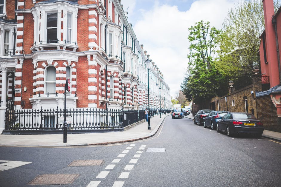 A street view of a residential area in Earls Court, showing a row of Victorian-style terraced houses with red and white brick facades, bay windows, and decorative cornices. The foreground features a narrow road with parked cars, including a black convertible and a silver sedan, lined along the curb. The pavement on the left side is partially visible, with a black wrought-iron fence surrounding a property. In the background, the street extends with additional parked vehicles and greenery from trees lining the sidewalk. The scene is captured during the daytime with natural lighting, illustrating a typical urban neighbourhood suitable for house removals and local moving services, as provided by Man and a Van Earls Court.